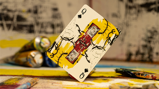 A queen of spades card featuring a Basquiat painting, placed on a table with various paint tubes.