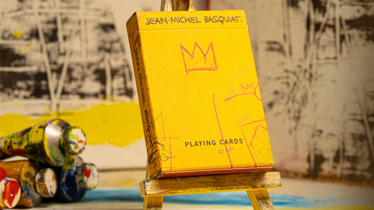A pack of playing cards with a yellow cover featuring a Basquiat painting, placed on a table with various paint cans and a wooden stand.