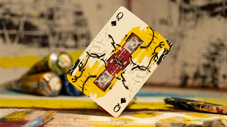 A queen of spades card featuring a Basquiat painting, placed on a table with various paint tubes.