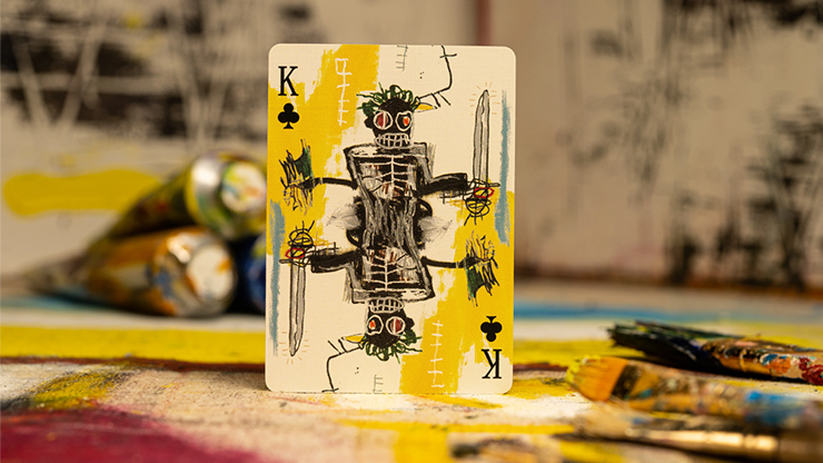 A King of clubs card featuring a Basquiat painting, placed on a table with various paint tubes.