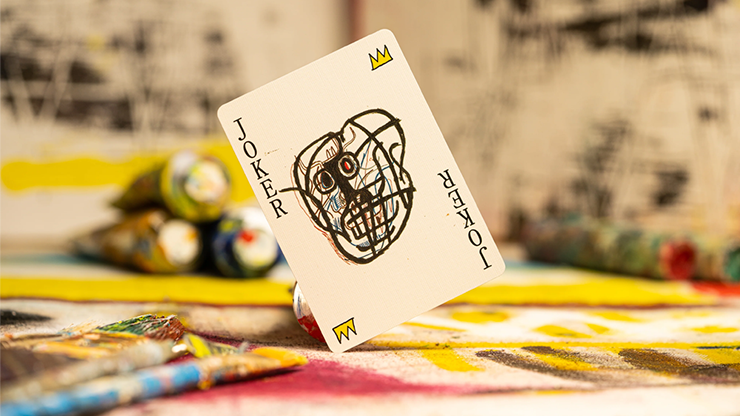A joker card featuring a Basquiat painting, placed on a table with various paint tubes.