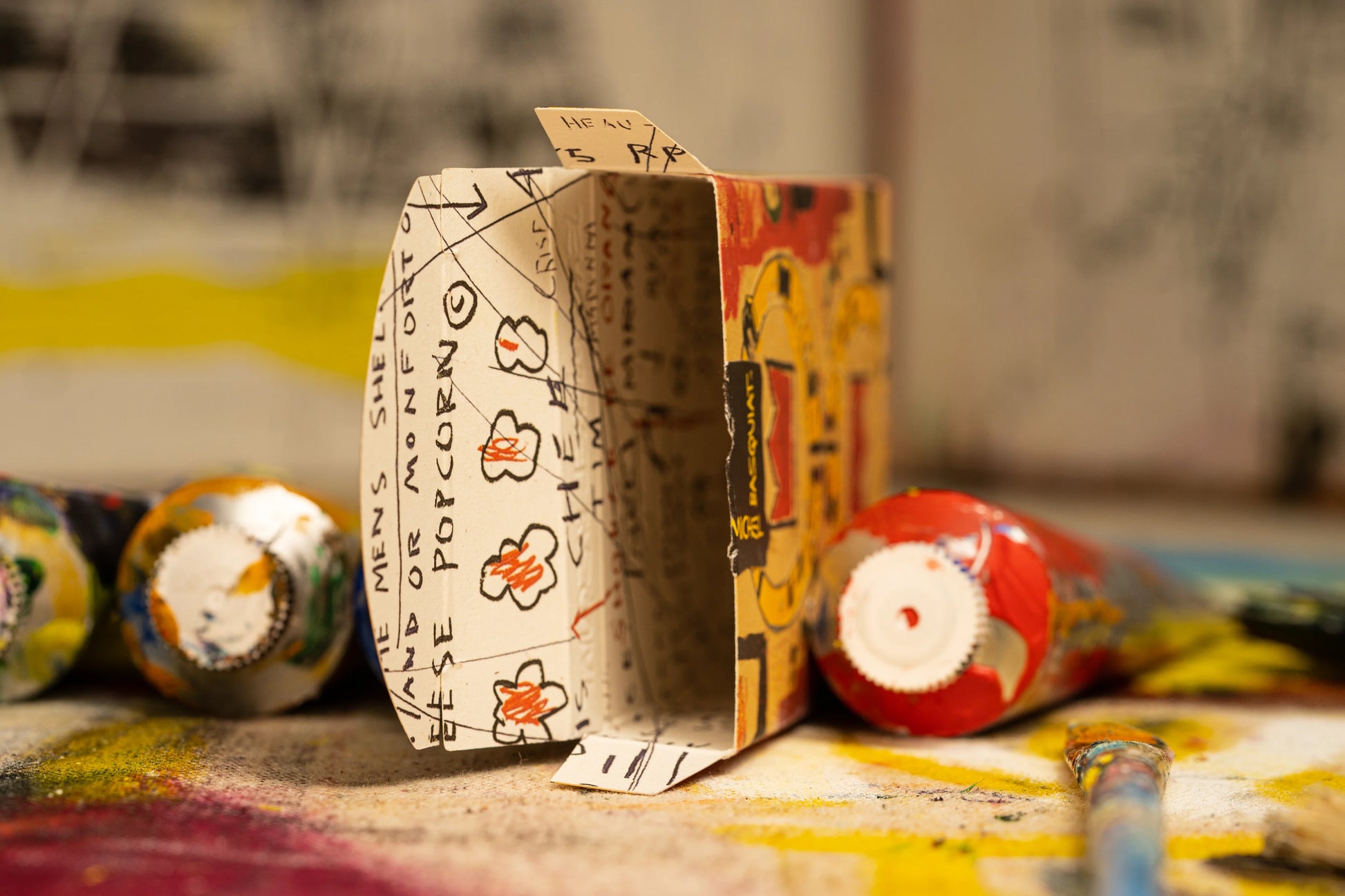 Tuck box interior features sketches of Jean-Michel Basquiat on a colorful background with tubes of paint.
